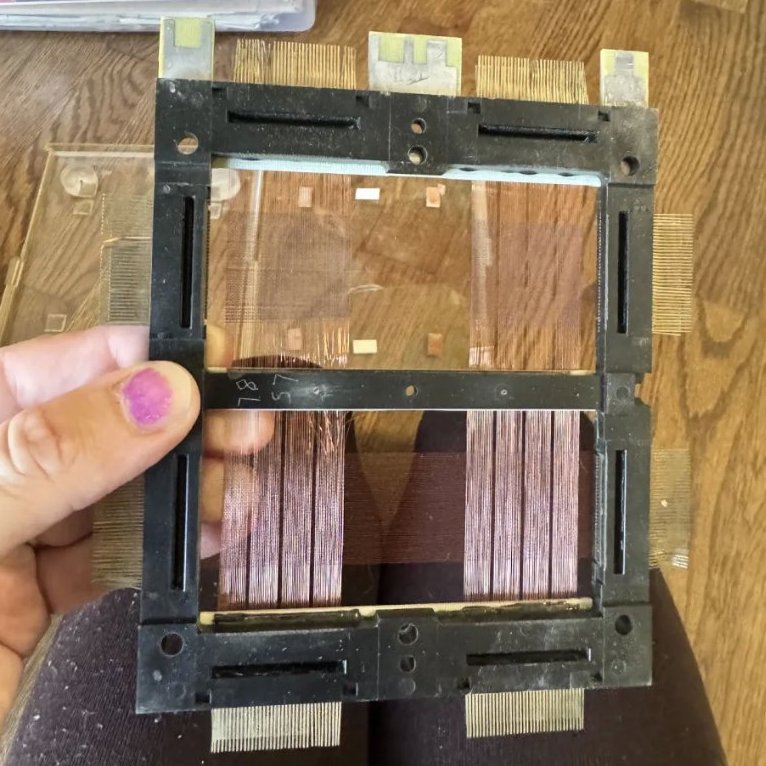A hand holding a magnetic core memory plane: a dark frame strung with fine copper wires crossing at tiny ferrite rings.